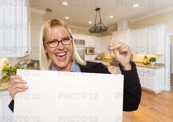 Happy young woman holding blank sign and keys inside beautiful custom kitchen