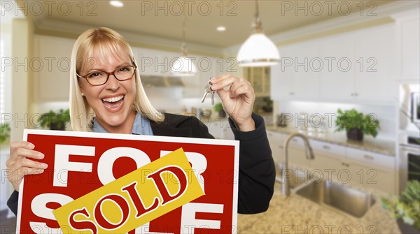 Happy young woman holding sold for sale real estate sign and keys inside beautiful custom kitchen