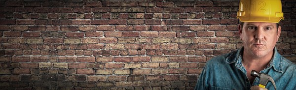 Male contractor in hard hat holding hammer in front of old brick wall banner with copy space