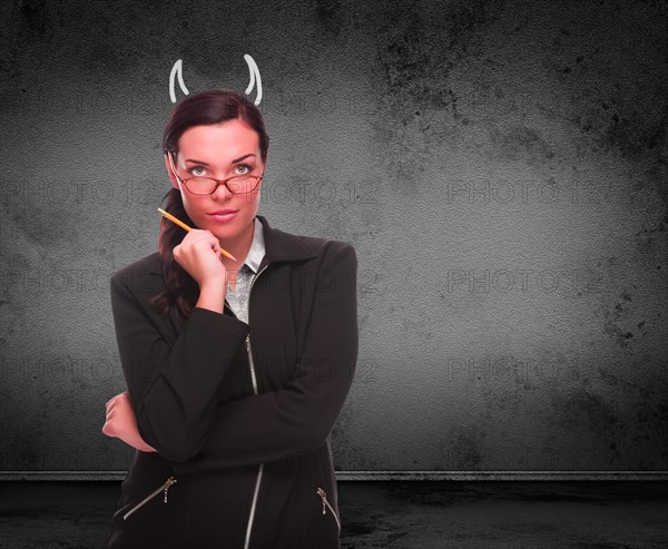 Devil horns drawn on head of red faced young adult woman with pencil in front of grungy wall with copy space