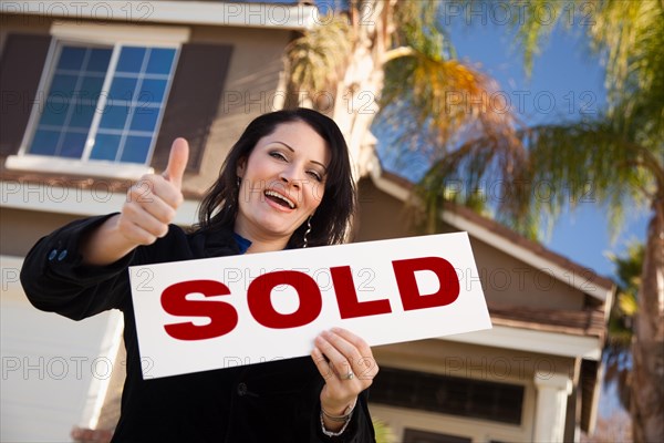 Happy attractive hispanic woman with thumbs up holding sold sign in front of house