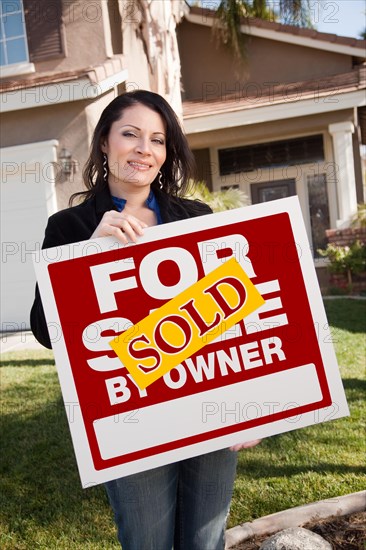 Happy attractive hispanic woman holding sold for sale by owner real estate sign in front of house