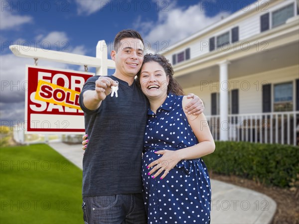 Happy hispanic couple in front of new home and sold real estate sign showing off their house keys