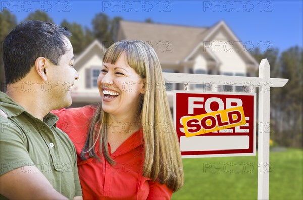 Happy mixed-race couple in front of sold home for sale real estate sign and house