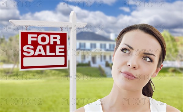 Thoughtful pretty mixed-race woman in front of home and house for sale real estate sign looking up and to the side