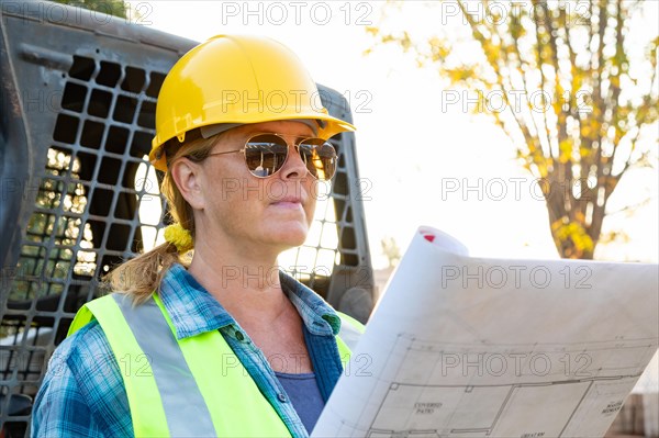 Female worker holding technical blueprints near small bulldozer at construction site