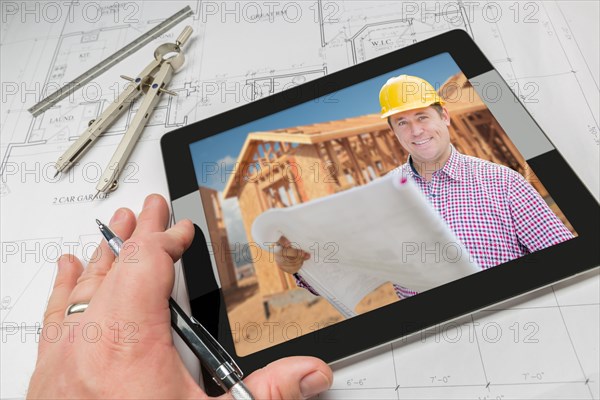Hand of architect on computer tablet showing contractor and home framing photo over house plans