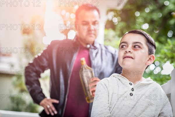 Afraid and bruised mixed-race boy in front of angry man holding bottle of alcohol