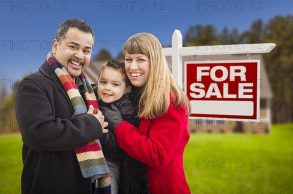 Warmly dressed young mixed-race family in front of home for sale real estate sign and house