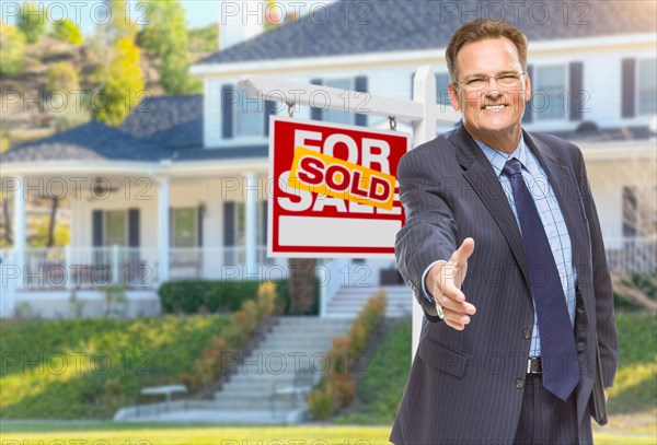 Smiling male agent reaching for a hand shake in front of beautiful house and sold real estate sign