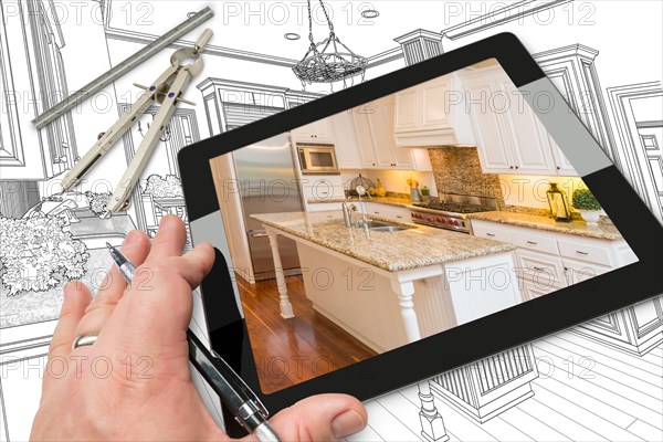 Hand of architect on computer tablet showing photo of kitchen drawing behind with compass and ruler