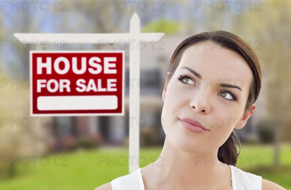 Thoughtful pretty mixed-race woman in front of home and house for sale real estate sign looking up and to the side