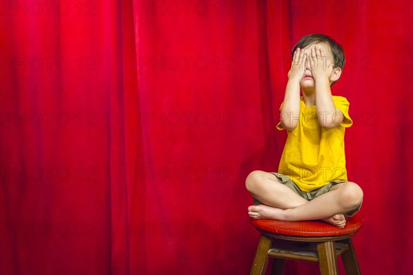 mixed-race boy boy covering his eyes sitting on stool in front of red curtain