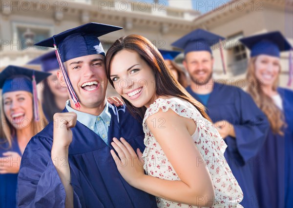 Proud male graduate in cap and gown with girl among other graduates behind