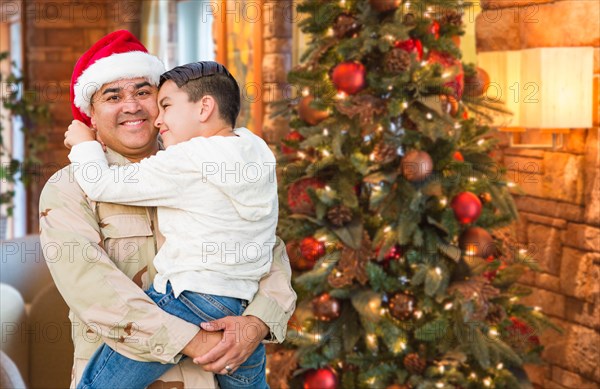 Hispanic armed forces soldier wearing santa hat hugging son in front christmas tree