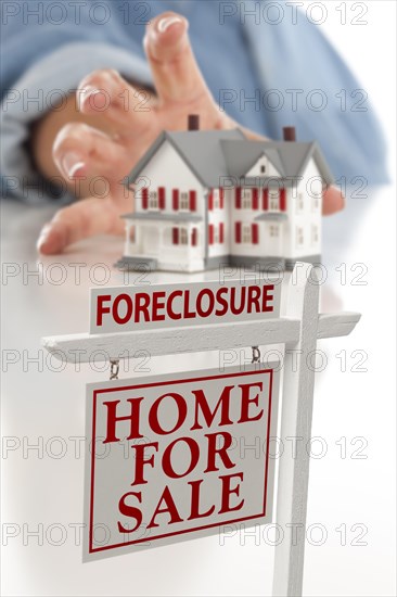 Foreclosure real estate sign in front of womans hand reaching for model house on a white surface