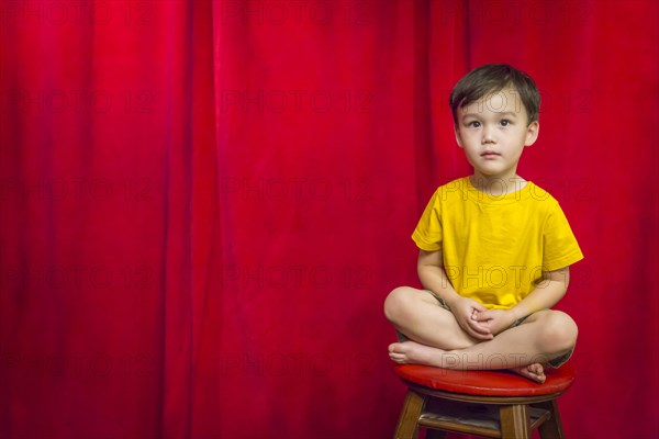 Handsome mixed-race boy sitting on stool in front of red curtain