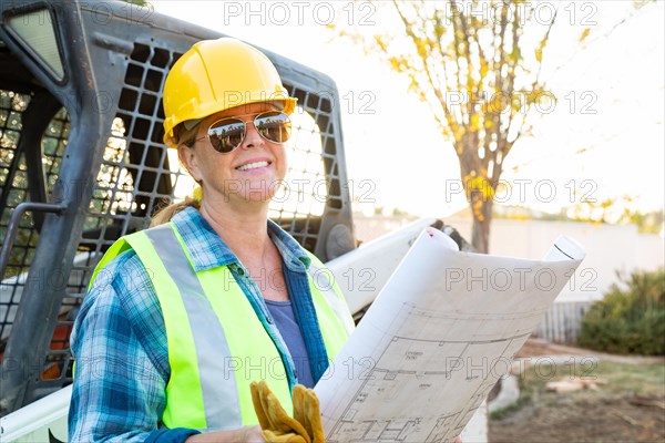 Smiling female worker holding technical blueprints near small bulldozer at construction site