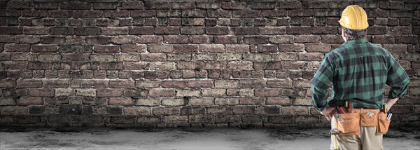 Contractor wearing hard hat and tool belt facing blank old brick wall banner with copy space