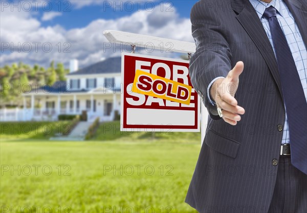 Real estate agent reaches for handshake with sold sign and new house behind