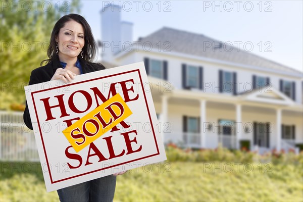 Smiling hispanic woman holding sold home for sale sign in front of beautiful house