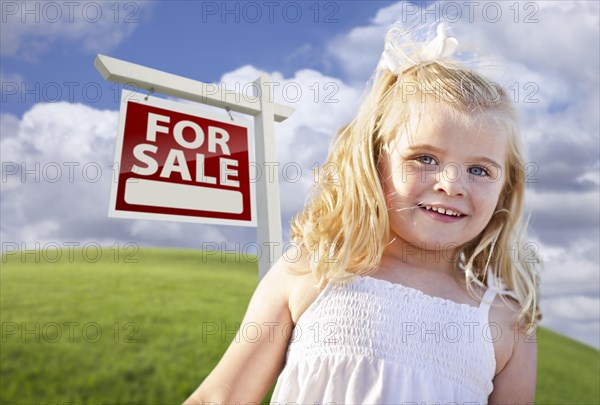 Adorable smiling girl in grass field with for sale real estate sign behind her