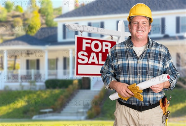 Contractor with plans and hard hat in front of for sale real estate sign and house