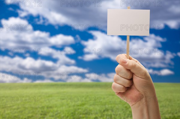 Blank sign in male fist over grass field and sky with clouds