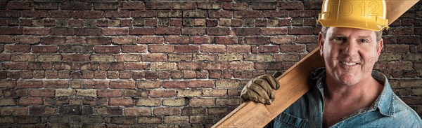 Male contractor in hard hat holding wood plank in front of old brick wall banner with copy space
