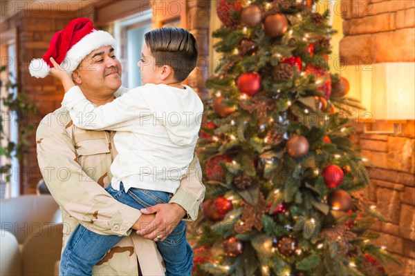 Hispanic armed forces soldier wearing santa hat hugging son in front christmas tree