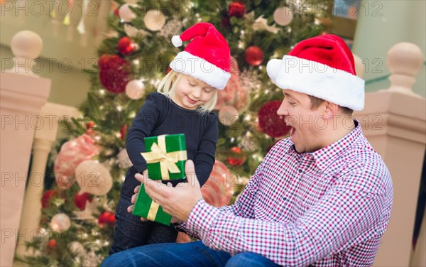 Happy young girl and father wearing santa hats opening gift box in front of decorated christmas tree