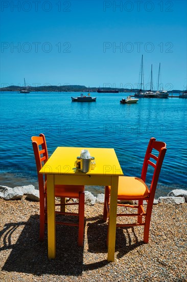 Yellow cafe restaurant table of street cafe with chairs on beach in Adamantas town on Milos island with Aegean sea with boats and yachts in background. Milos island