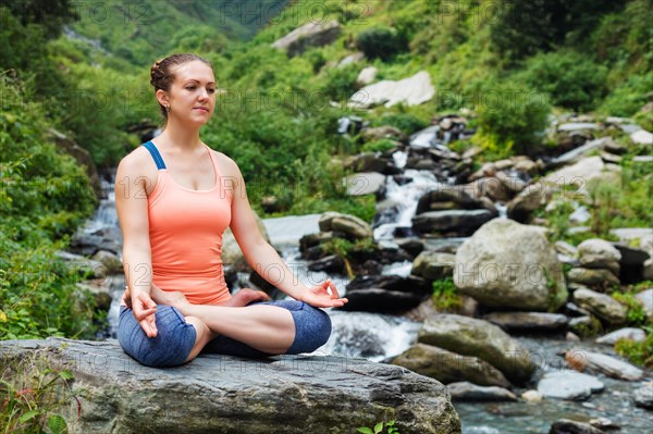 Woman in Hatha yoga asana Padmasana outdoors at tropical waterfall