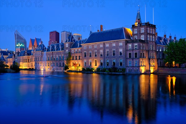 View of the Binnenhof House of Parliament and the Hofvijver lake with downtown skyscrapers in background illuminated in the evening. The Hague