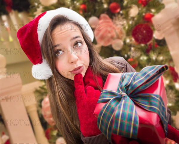 Thinking girl wearing A christmas santa hat with bow wrapped gift in front of decorated tree