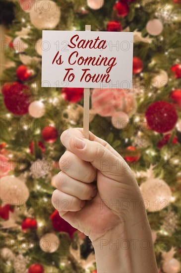 Hand holding santa is coming to town card in front of decorated christmas tree