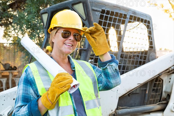Smiling female worker holding technical blueprints near small bulldozer at construction site