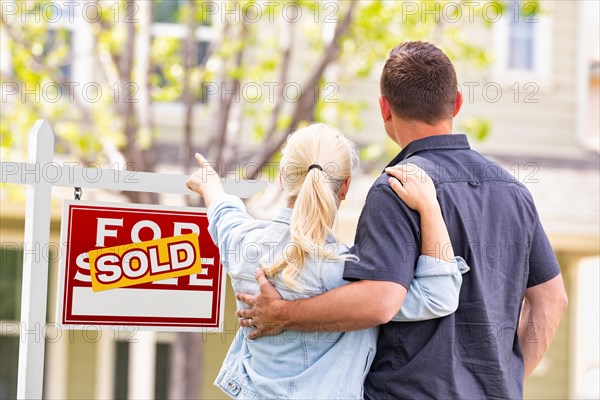 Caucasian couple facing and pointing to front of sold real estate sign and house