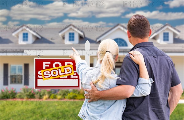 Young adult couple facing and pointing to front of sold real estate sign and house