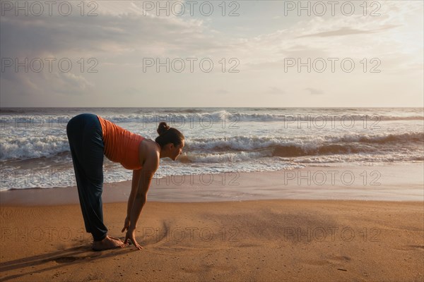 Young sporty fit woman doing yoga Sun salutation Surya Namaskar pose Uttanasana on tropical beach on sunset. With lens flare