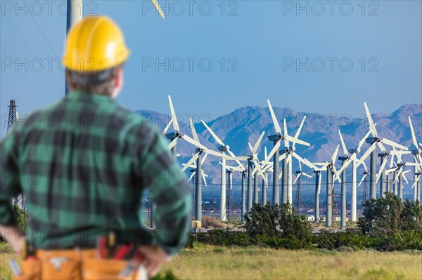 Male contractor wearing tool belt and hard hat facing alternative energy wind turbines