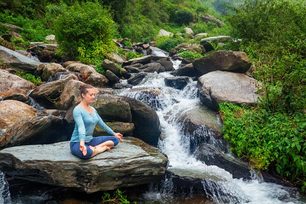 Woman in Hatha yoga asana Padmasana outdoors at tropical waterfall