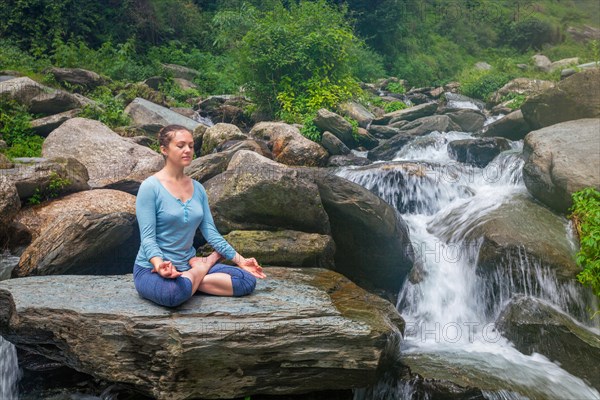 Woman in Hatha yoga asana Padmasana outdoors at tropical waterfall