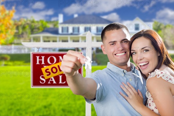 mixed-race excited military couple in front of new home with new house keys and sold real estate sign outside