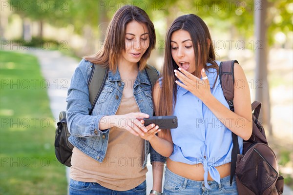 Two beautiful young ethnic twin sisters with backpacks using A smartphone outside