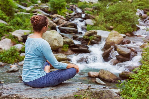 Woman in Hatha yoga asana Padmasana outdoors at tropical waterfall