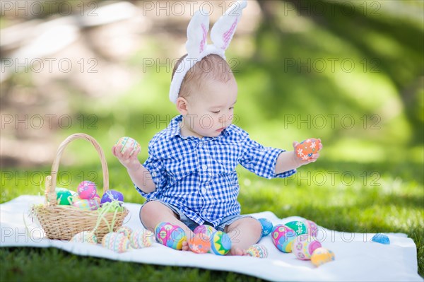 mixed-race chinese and caucasian baby boy outside wearing rabbit ears playing with easter eggs