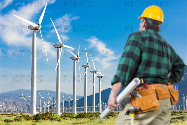 Male contractor wearing tool belt and hard hat facing alternative energy wind turbines