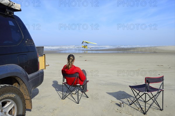 Woman sitting on camping chair next to all-terrain vehicle on beach watching a light aircraft taking off