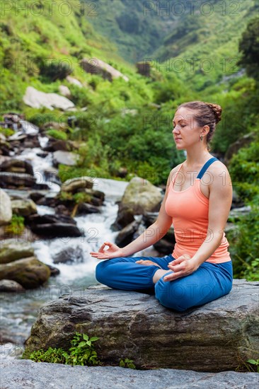 Woman in Hatha yoga asana Padmasana outdoors at tropical waterfall